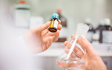 Eine Hand hält ein Behälter mit Pillen, während die andere Hand eine durchsichtige Flasche mit einer Flüssigkeit hält.