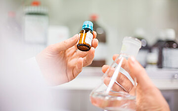 Eine Hand h&auml;lt ein Beh&auml;lter mit Pillen, w&auml;hrend die andere Hand eine durchsichtige Flasche mit einer Fl&uuml;ssigkeit h&auml;lt.