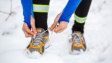 Das Bild zeigt F&uuml;&szlig;e und Beine unterhalb des Knies auf Echneeunterlage und zwei H&auml;nde, die den rechten Joggingschuh schn&uuml;ren.