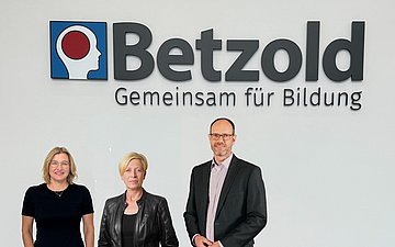 Bezirksratsmitglieder der AOK Ostwürttemberg bei Betzold Tina Betzold steht mit Heike Madan und Markus Kilian vom Bezirksrat der AOK Ostwürttemberg vor einem Betzold-Logo.