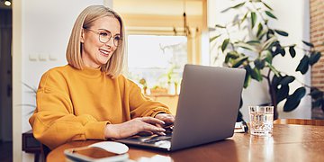 Frau vor dem Laptop Das Bild zeigt eine blonde Frau mit Brille am Küchentisch vor einem Laptop bei der Recherche. Sie trägt einen dunkelgelben Pullover. Rechts neben dem Laptop steht ein Glas mit Wasser.