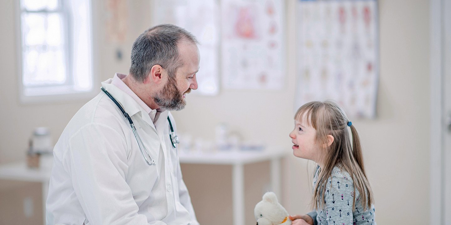 Doktor sitzte neben einem Mädchen im Behandlungszimmer. Das Kind hält einen Teddybär.