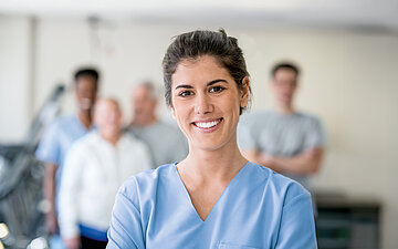 Eine junge Frau in blauer Pflegekleidung l&auml;chelt in die Kamera.