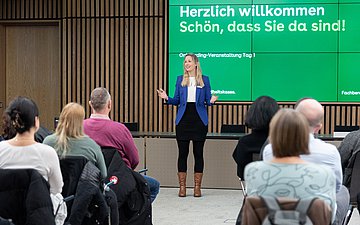 In einem mit Holz vertäfelten Saal sitzen zahlreiche Männer und Frauen und blicken auf eine kleine Bühne. Dort wird eine Begrüßung der Versammelten an die Wand projiziert.