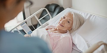 Eine ältere Dame liegt auf einem Krankenhausbett und schaut eine Pflegerin an.
