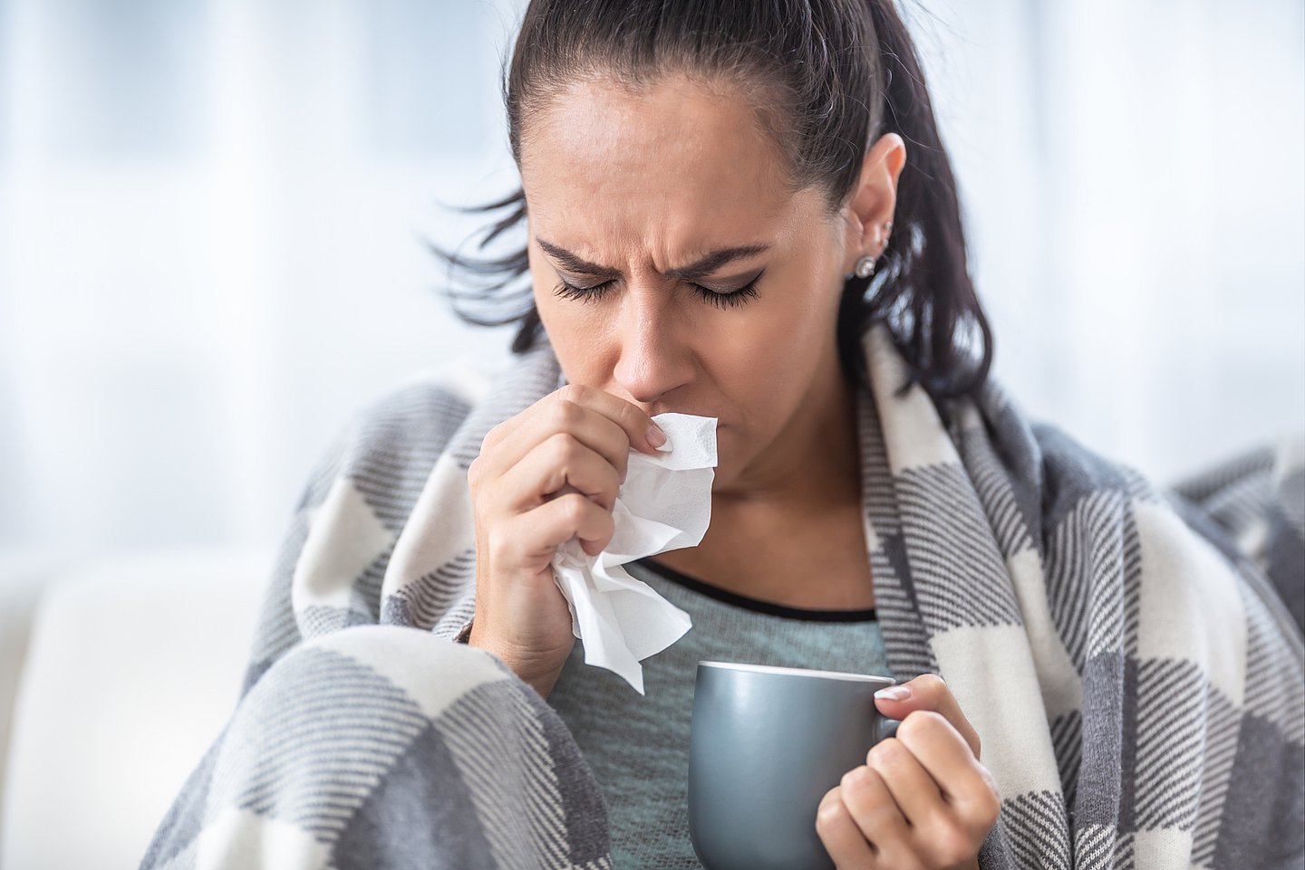 Foto zeigt eine Frau mit Infekt, die sich mit der einen Hand ein Papiertaschentuch vor die Nase hält, in der anderen hält sie eine Tasse und um die Schultern liegt eine warme Decke.