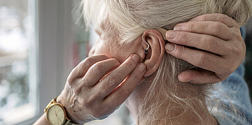 Eine &auml;ltere Frau packt sich ein H&ouml;rger&auml;t ins linke Ohr.