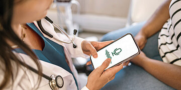 Eine &Auml;rztin sitzt einer Patientin gegen&uuml;ber und h&ouml;rt zu. In ihrer Hand h&auml;lt sie ein Smartphone, auf dessen Bildschirm ein Mikrofon-Symbol zu sehen ist.