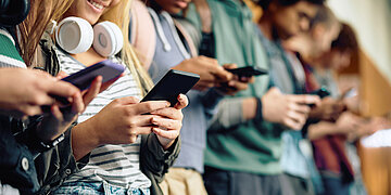 Mehrere Jugendliche stehen in einer Reihe nebeneinander. Alle sind am Smartphone.