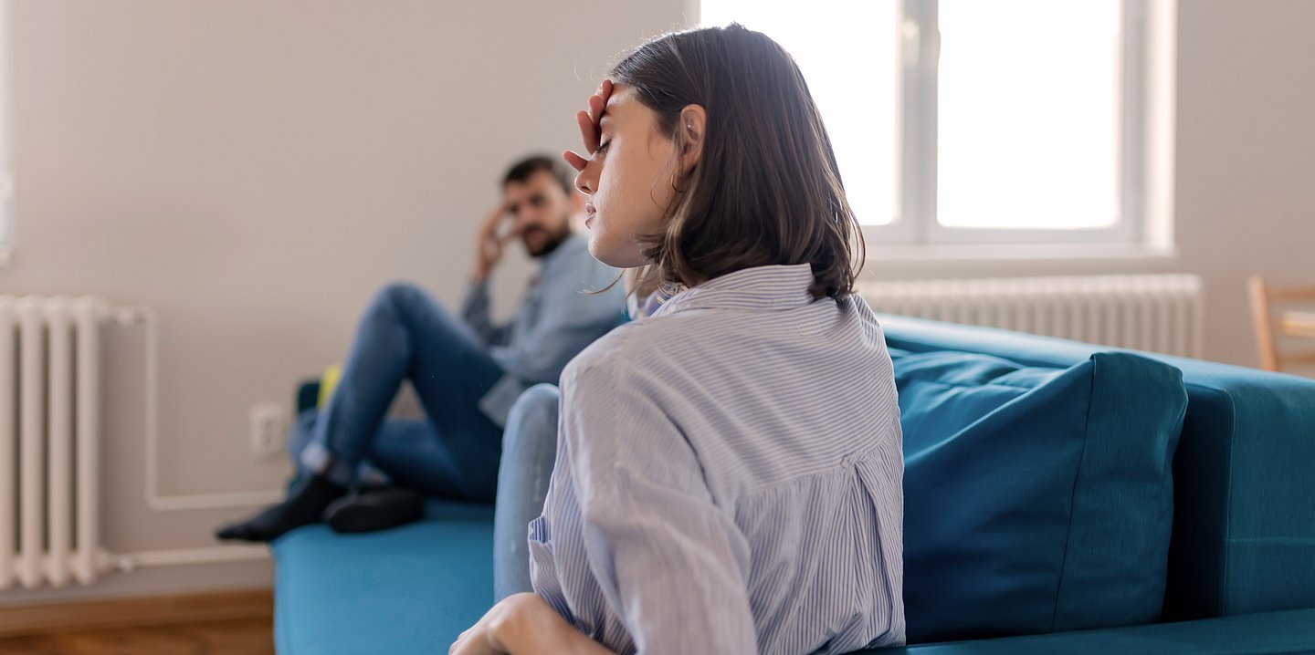 Foto: Ein Mann und eine Frau sitzen in Abstand zueinander auf einer Couch. Beide greifen sich an die Stirn und schauen traurig.