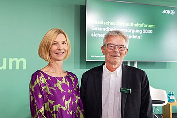 Das Bild zeigt eine blonde Frau (links) in violetter Bluse mit hellgr&uuml;nen Farbflecken und einen graumelierten Brillentr&auml;ger mit dunkelgrauem zart karierten Sakko und offenem wei&szlig;en Hemd.