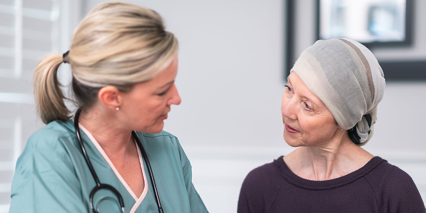 Foto: Eine Frau in Medizinerkleidung spricht mit einer Frau, die ein Tuch über dem Kopf trägt.