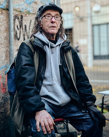 Ein älterer Mann sitzt mit Schiebermütze und Rucksack auf einem Stuhl und schaut in die Ferne. 