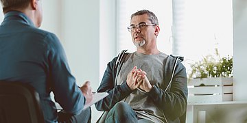 Unscharf im Vordergrund sitzt ein Mann mit Klemmbrett. Vor ihm sitzt ein mittelalter Mann, der mit dem Therapeuten redet.