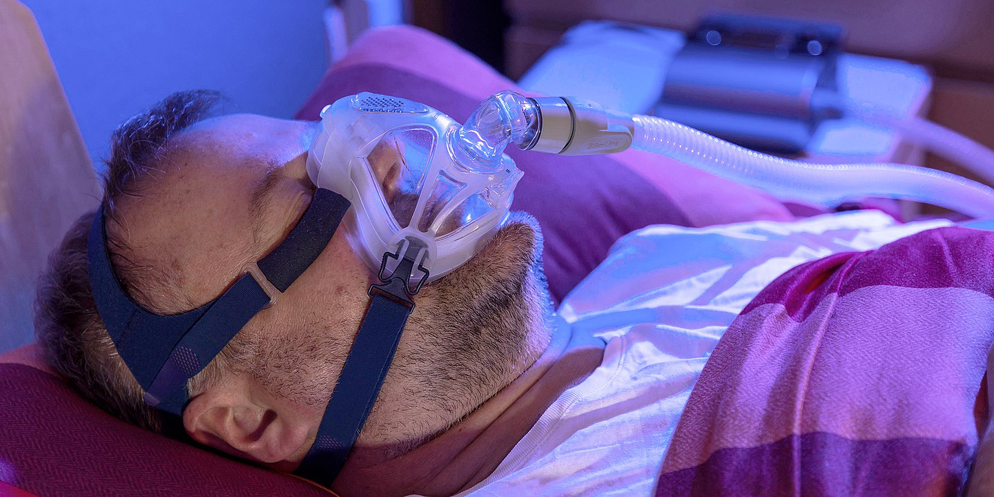 Foto: Ein Mann liegt im Bett und trägt eine Maske, die seine Atmung unterstützt.