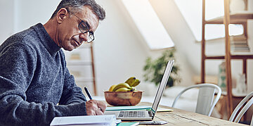 Foto: Ein Mann sitzt am E&szlig;tisch und notiert etwas, vor ihm ein Laptop.