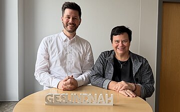 Pascal Haggenm&uuml;ller und Petra Spitzm&uuml;ller stehen gemeinsam an einem runden Tisch, auf dem eine Kleinplastik das Wort "gesundnah" zeigt.