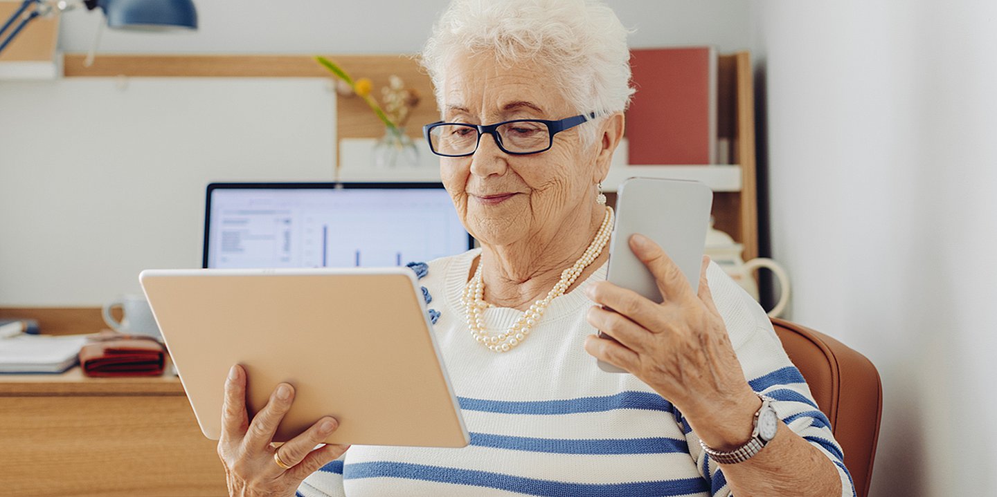 Foto: Eine ältere Frau hält in der einen Hand ein Tablet, in der anderen ein Smartphone. Im Hintergrund steht ein Laptop.