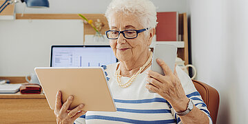 Foto: Eine &auml;ltere Frau h&auml;lt in der einen Hand ein Tablet, in der anderen ein Smartphone. Im Hintergrund steht ein Laptop.