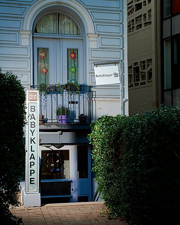 Babyklappe Zu sehen ist die Babyklappe in Hamburg-Altona. Ein Altbau-Gebäude mit absteigenden Treppen zum Eingang und zur Klappe. Darüber ein französischer Balkon, die Fenster geschmückt mit bunten Blumengirlanden. Links ein großes Schild mit dem Wort "Babyklappe".