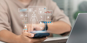 Im Hintergrund sitzt eine Person an einem Laptop, schaut aber auf ein Smartphone. Da&uuml;ber schwebt sozusagen ein Hologramm von einem Menschen, neben dem Gesundheits-Icons und Werte angezeigt werden.