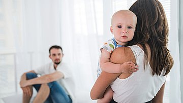 Wochenbettdepression Väter Eine Frau hält ein kleines, weinendes Kind auf dem Arm, ein Mann sitzt mit verschränkten Armen auf dem Boden.