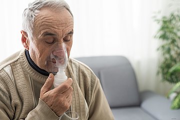 Ein älterer Mann sitzt auf einem Sofa und inhaliert mit einem Inhalationsgerät.