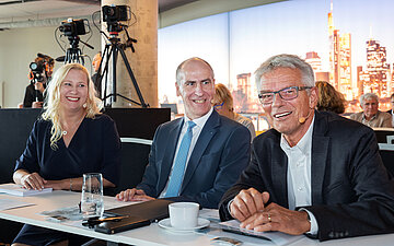 Das Bild zeigt von links nach rechts eine blonde Frau mit langen Haaren und dunkelblauem Kelid, einen Mann mit Halbglatze stahlblauem Sakko, wei&szlig;em Hemd und hell-dunkelblau-karrierter Krwatte, einen Mann mit vollem grau-melierten Haar, blau-karierten Sakko und offenem wei&szlig;en Hemd., 