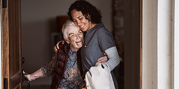 Eine Pflegerin im Arbeitskittel umarmt eine &auml;ltere Frau. Beide l&auml;cheln.