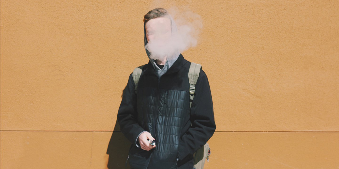 Foto: Junger Mann mit E-Zigarette in der Hand – sein Gesicht ist von Rauch verdeckt.