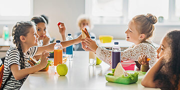 Foto: Mehrere Kinder sitzen gemeinsam am Tisch und teilen sich in einer Schule ihre Pausenbrote und Getr&auml;nke.