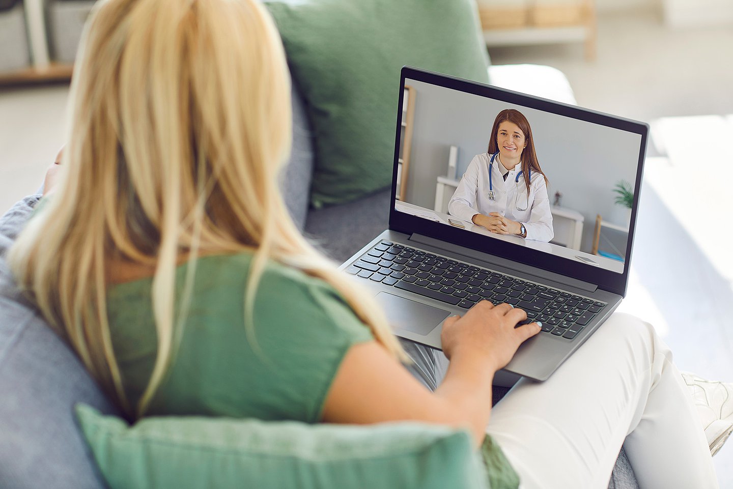 Eine weibliche Patientin sitzt auf in ihrem Wohnzimmer auf einer Couch und spricht per Videosprechstunde mit ihrer behandelnden Ärztin, die auf dem Bildschirm des Laptops sichtbar ist.