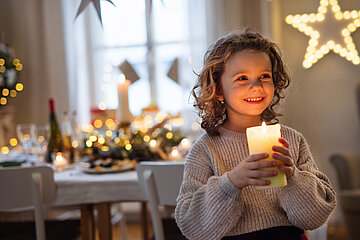 Ein kleines M&auml;dchen steht mit einer brennenden Kerze in der Hand im weihnachtlich geschm&uuml;ckten Wohnzimmer.