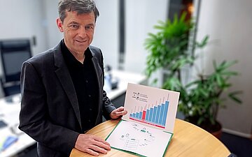 Hans-Joachim Seuferlein steht an einem Tisch und zeigt ein Diagramm zur HZV.