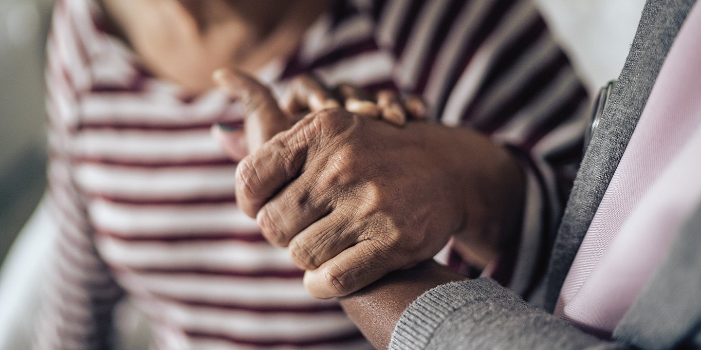 Foto einer alten Dame, die von einer Hand gestützt wird. Die Gesichert sind nicht zu sehen.