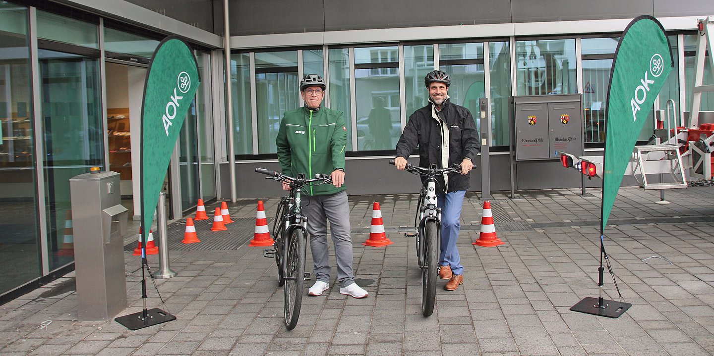 Foto zeigt zwei Männer mit Helm und Fahrrad vor einem Verwaltungsgebäude