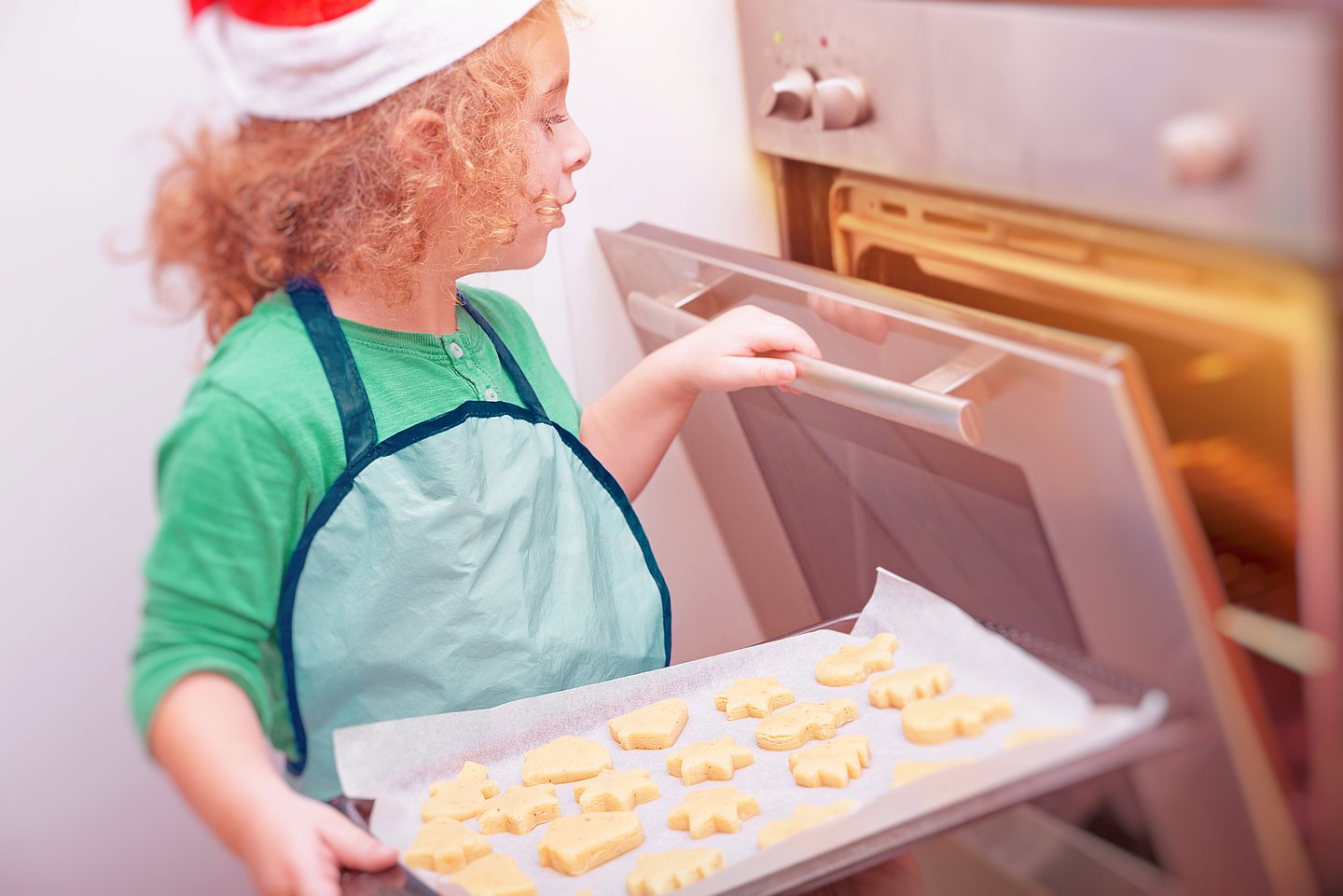 Foto: Ein kleines Mädchen mit Nikolausmütze steht am Backofen und will ein Blech mit ausgestochenen Keksen in den Backofen schieben.