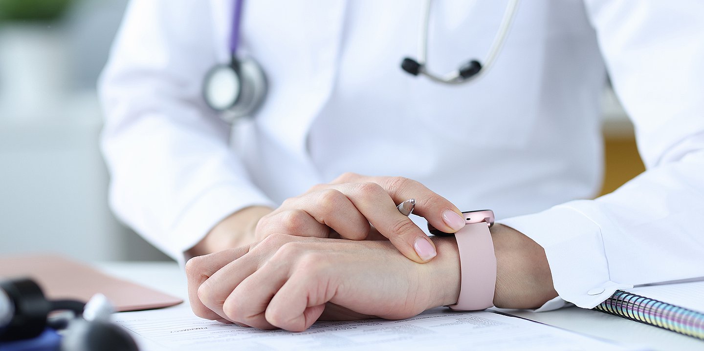 Foto einer Person im medizinischen Kittel mit umgehängtem Stethoskop, die auf eine Armbanduhr blickt. Das Gesicht ist nicht zu sehen.