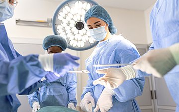 In einem OP-Saal stehen mehrere Chirurginnen und Hilfsärztinnen um den OP-Tisch, operieren und reichen sich Instrumente.