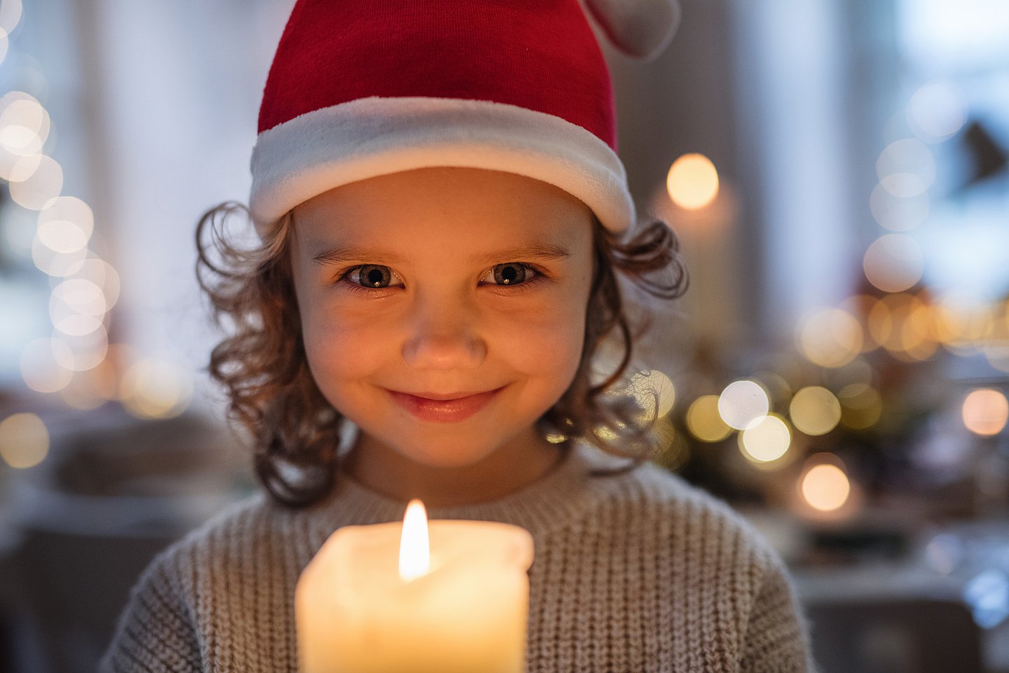 Ein kleines Mädchen, das eine rote Nikolausmütze auf dem Kopf trägt, steht vor einer brennenden Kerze oder hält diese in der Hand. Sie freut sich und schaut lächelnd in die Kamera.