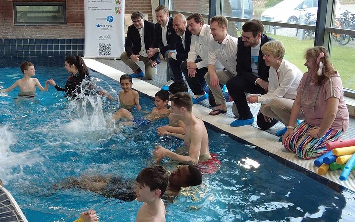 Foto zeigt Kinder im Wasserbecken bei Schwimmübungen. Schulministerin Feller und die Kooperationspartner des Programms "NRW kann schwimmen!" schauen ihnen am Beckenrand dabei zu.