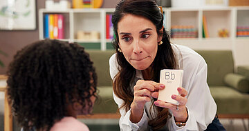 Eine Logopädin übt mit einem Mädchen, das von hinten gezeigt wird, den Buchstaben 'B'.