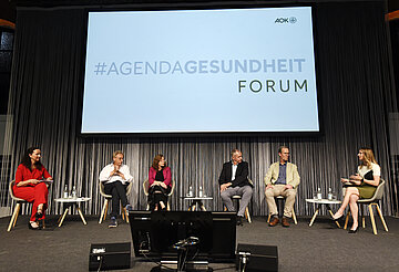 Podiumsg&auml;ste sitzen vor einem gro&szlig;en Bildschirm: Daniela Wiesler (Moderation), Prof. Dr. Alexander Woll, Ministerialdirektorin Leonie Dirks, Prof. Dr. Mark Dominik Alscher, Prof. Dr. Hajo Zeeb, Gordana Marsic