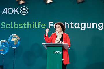 Dr. Carola Reimann steht vor einer gr&uuml;nen Stellwand mit der Aufschrift "Selbsthilfe-Fachtagung" hinter einem Stehpult und h&auml;lt eine Rede.