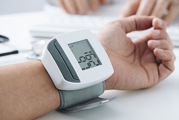 Eine linke Hand und Unterarm liegen locker auf einem Tisch. Die Handinnenfläche zeigt nach oben, am Handgelenk ist ein digitales Blutdruckmessgerät angebracht, dass einen bestimmten Wert anzeigt.