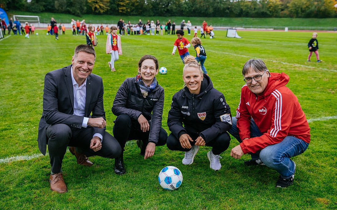 Vier Personen positionieren sich mit einem Ball auf dem Rasen eines Fußballstadions.