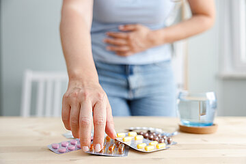 Eine Frau steht an einem Tisch, sie fasst sich mit der linken Hand an ihren Magen, w&auml;hrend sie mit der rechten nach einem der Tablettenblister greift, die auf dem Tisch vor ihr liegen. Daneben steht ein Glas Wasser.