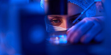 Close Up einer Person mit medizinischer Sicherheitskleidung in Blau. Haube, Mundschutz und Handschuhe. Zu sehen ist lediglich die Augenpartie.