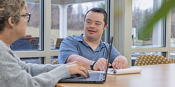 Ein Mann mit Behinderung bekommt von einer Frau etwas am Laptop gezeigt.