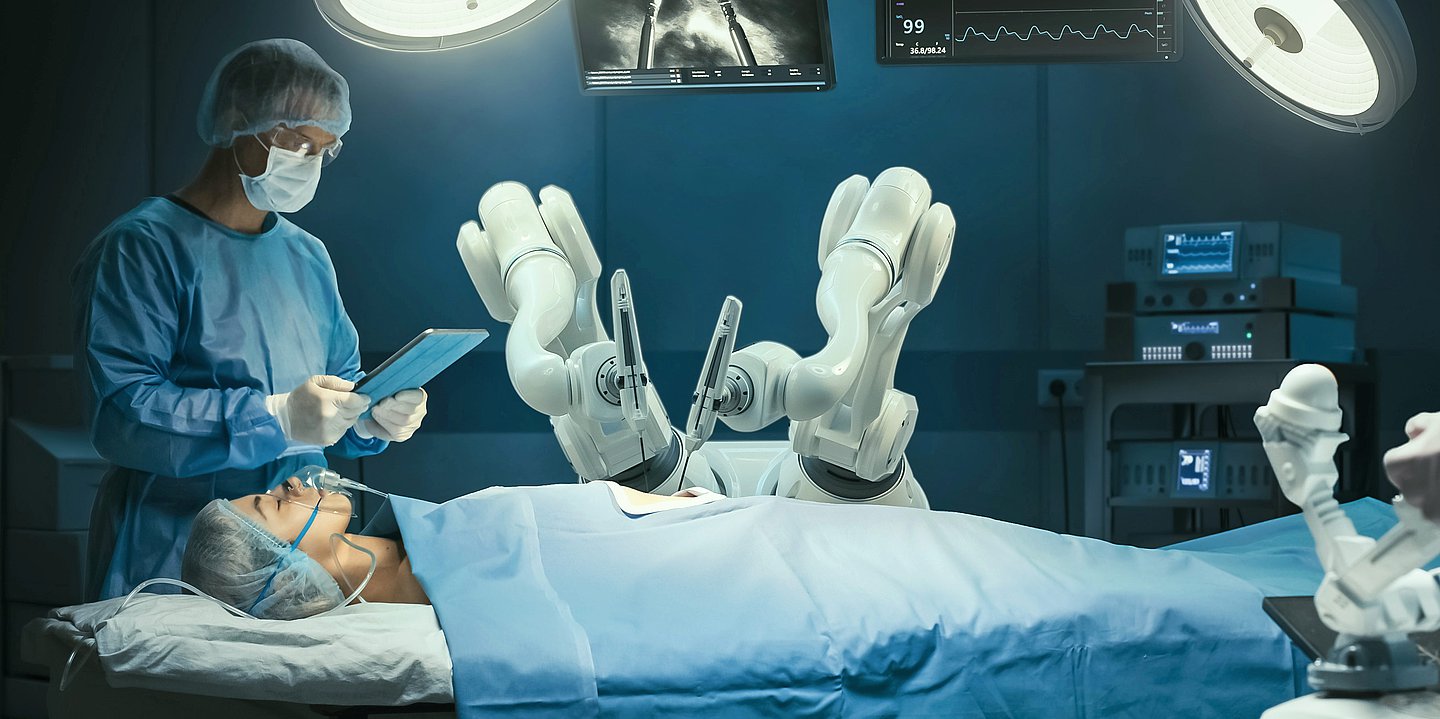 Foto: In einem Operationsaal schaut ein Mann in OP-Kleidung auf ein Tablet während ein Patient auf dem OP-Tisch liegt und Robotergreifarme über ihm schweben.